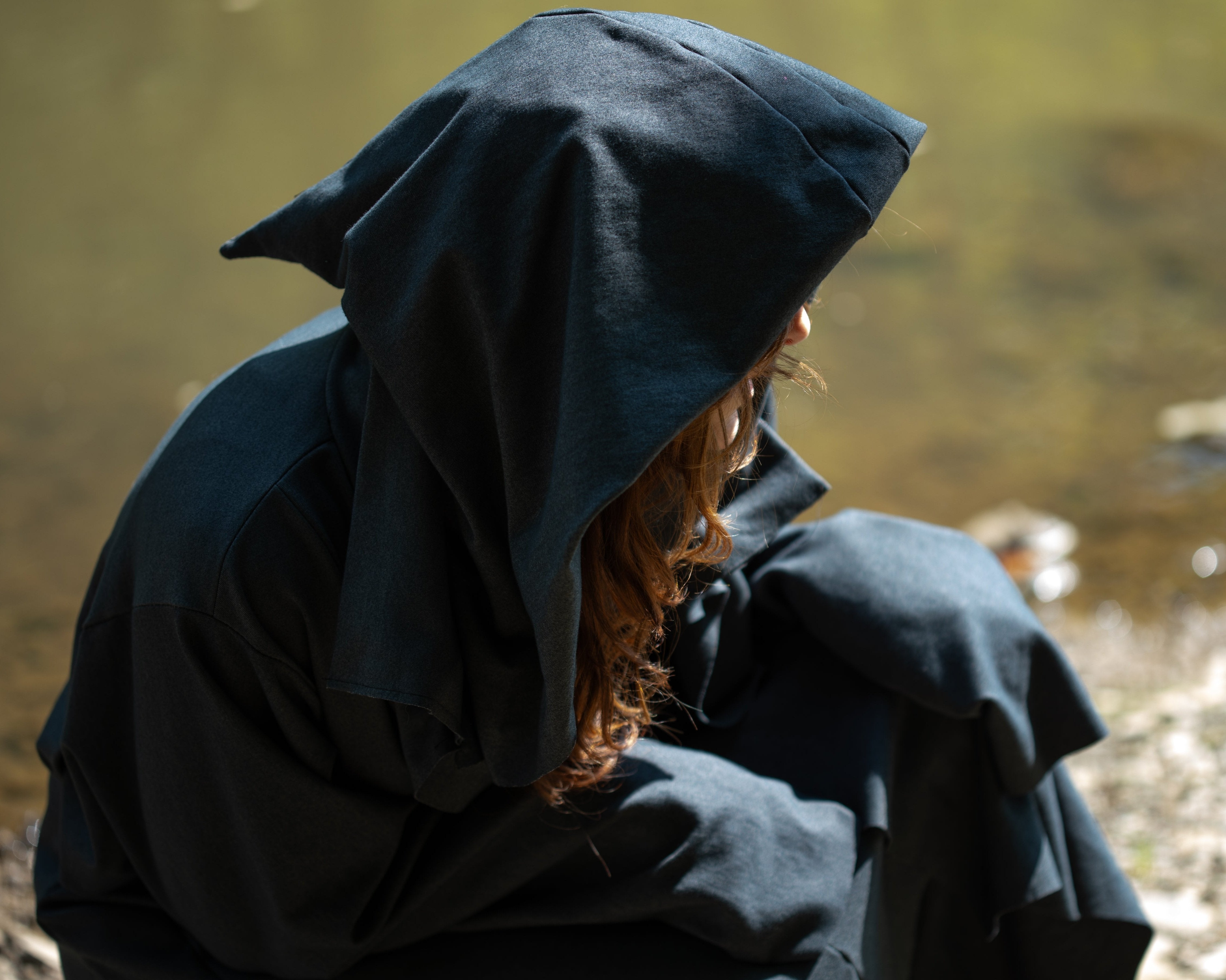 Person wearing a long hooded coat with a hood by a river bank