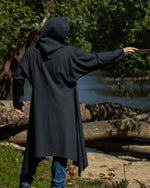 back view of a person wearing a long hooded coat standing by a river