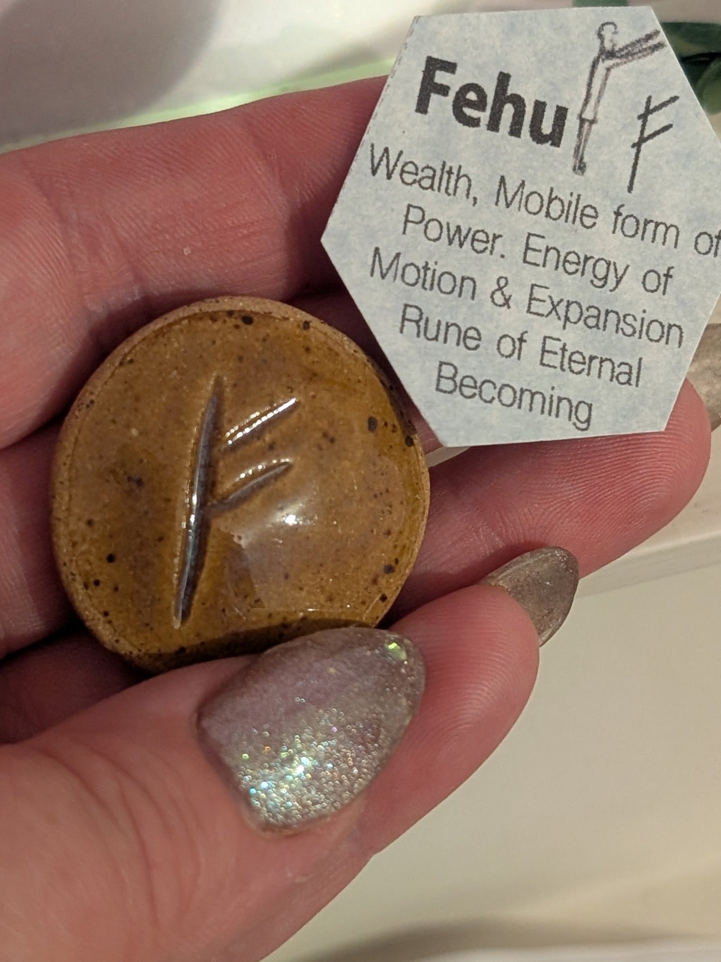 ceramic disc with a rune and a label describing 'Fehu' rune held in a hand.